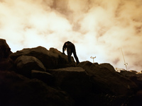 A migrant's body is silhouetted against the bright clouds bouncing light from the port's light posts as he climbs the rock that separate the...