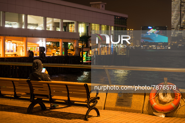 A man checks his phone as he sits by himself near the Central Piers. In the background, the M+ museum's facade can be seen, in Hong Kong, Ch...