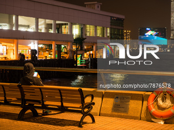 A man checks his phone as he sits by himself near the Central Piers. In the background, the M+ museum's facade can be seen, in Hong Kong, Ch...