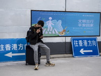 A man sitting on a suitcase next to signs pointing to Hong Kong at Shenzhen Bay Port on January 8, 2023 in Shenzhen, China. Hong Kong today...